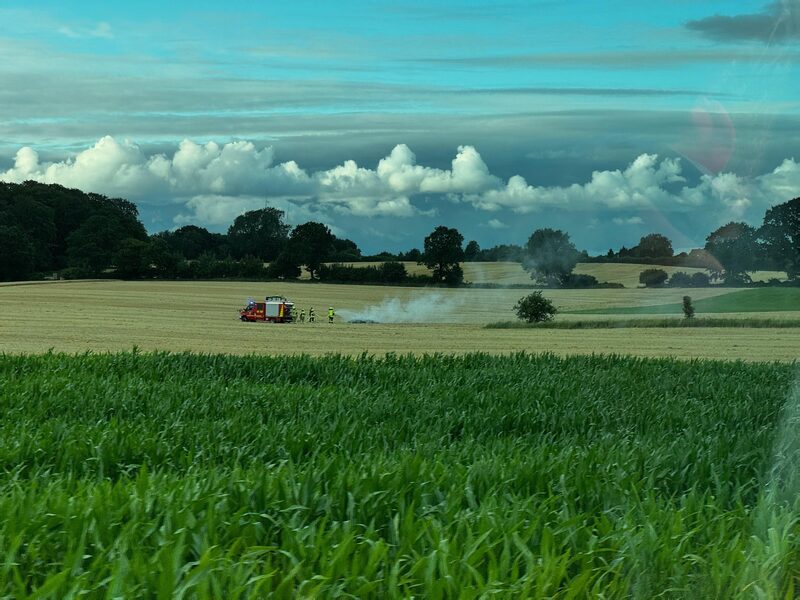 Weites Feld, am Horizont steigt Rauch auf, daneben Einsatzfahrzeuge.