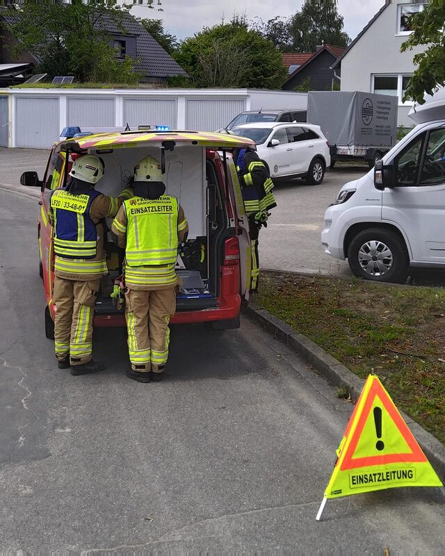 Zwei Einsatzkräfte in gelben Warnwesten mit der Aufschrift „Einsatzleiter“ am offenen Heck eines roten Einsatzleitwagens. Vorne steht ein Warndreieck „Einsatzleitung“.
