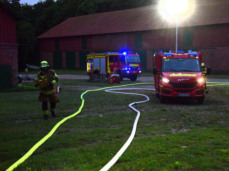 Abendliche Übung: Schläuche liegen auf einer Wiese vor einer Scheune, zwei Löschfahrzeuge mit Blaulicht.