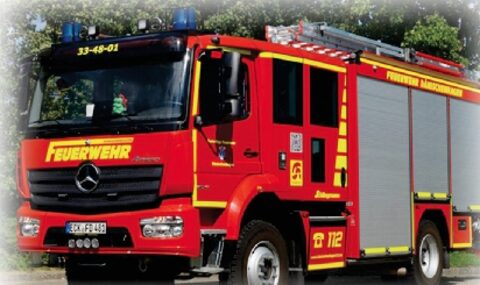 Rotes Löschgruppenfahrzeug mit gelben Streifen, Aufschrift „Feuerwehr“ und Blaulicht auf dem Dach, schräg von vorn.