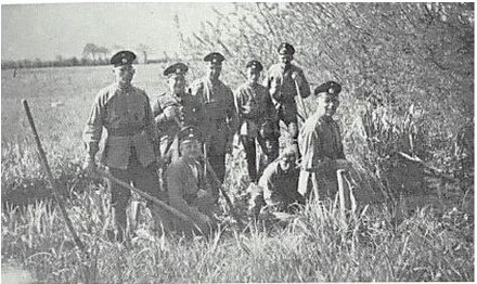 Altes Schwarz-Weiß-Foto: neun Männer in Uniform mit Werkzeug im hohen Gras.