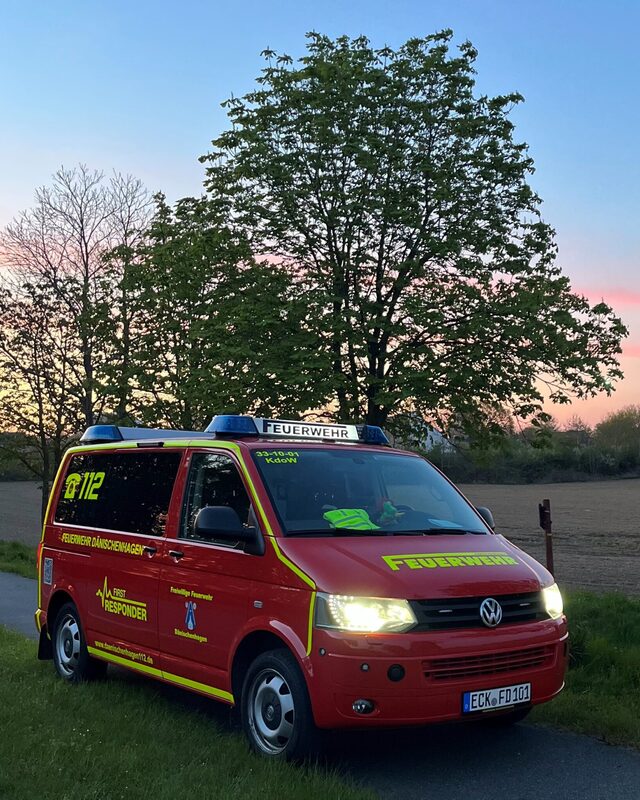 Der rote VW-Bus der First Responder mit eingeschaltetem Licht auf einem Feldweg, dahinter Bäume im Abendrot.