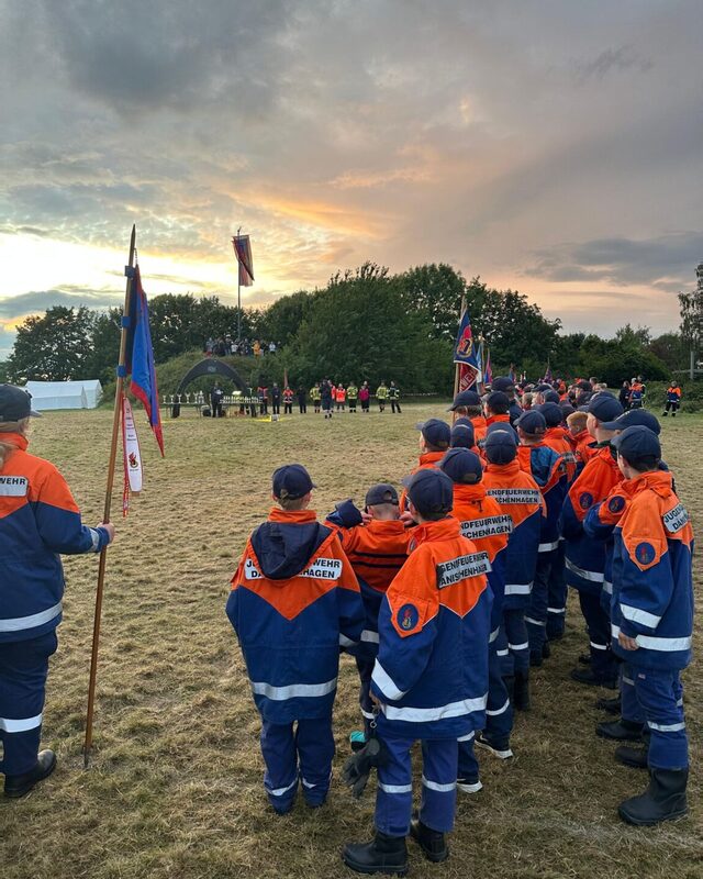 Jugendliche in blau-orangen Uniformen stehen in Reihen auf einer Wiese. Vorne wird eine Fahne gehalten, der Himmel färbt sich abendlich.