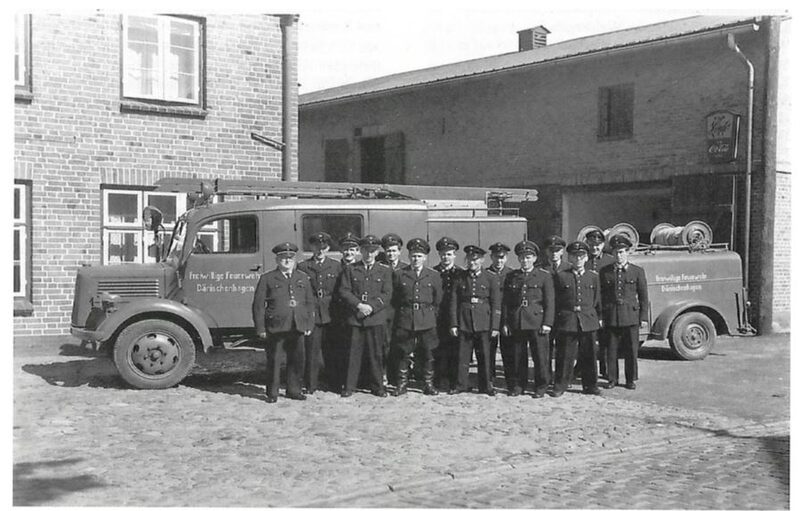 Schwarz-Weiß-Foto 1954: vierzehn Feuerwehrmänner in Uniform auf einem Kopfsteinpflasterhof zwischen einem alten Löschfahrzeug und einem Schlauchanhänger.