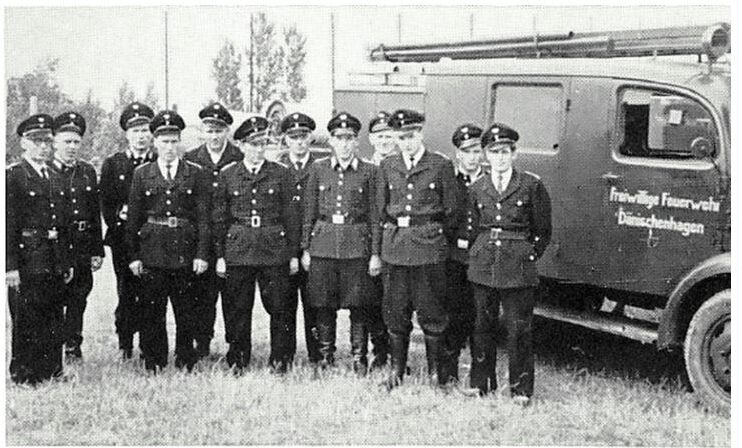 Schwarz-Weiß-Foto 1961: zwölf Feuerwehrmänner in Uniform mit Schirmmütze und Koppel neben einem Löschfahrzeug mit der Aufschrift „Freiwillige Feuerwehr Dänischenhagen“.