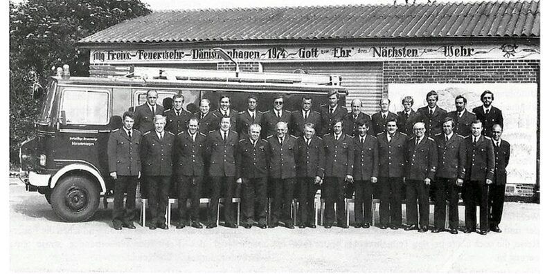 Schwarz-Weiß-Foto 1979: die Wehr in zwei Reihen vor einem Löschfahrzeug und dem alten Gerätehaus mit dem Schriftzug „1889 Freiw. Feuerwehr Dänischenhagen 1974 – Gott zur Ehr, dem Nächsten zur Wehr“.