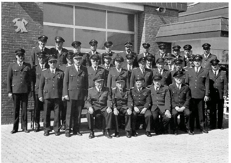 Schwarz-Weiß-Foto 1989: die Wehr in Uniform und Schirmmütze in drei Reihen vor dem Gerätehaus, vorne fünf ältere Kameraden auf Stühlen.