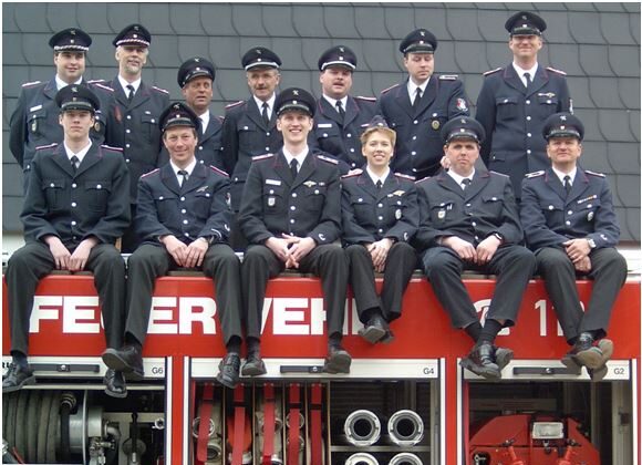 Gruppenfoto 2004: dreizehn Feuerwehrleute in Uniform und Schirmmütze sitzen und stehen auf dem Dach eines Löschfahrzeugs mit geöffneten Geräteräumen.