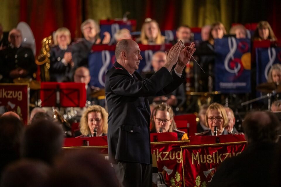 Ein Dirigent in dunkler Uniform hebt beide Hände. Hinter ihm spielt der Musikzug, vor den Notenständern hängen rote Banner mit der Aufschrift Musikzug.