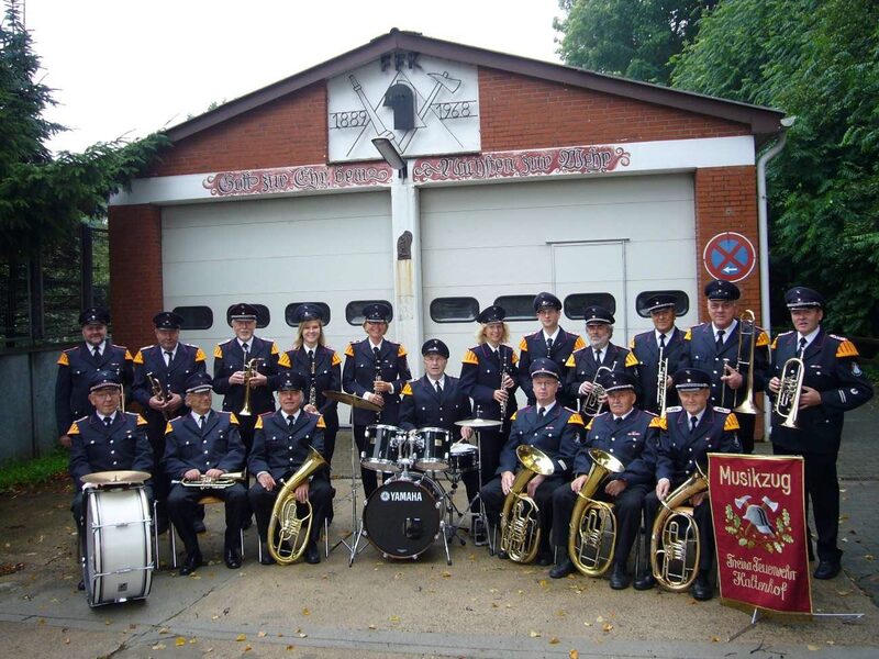 Gruppenfoto: rund zwanzig Musikerinnen und Musiker in dunkelblauer Uniform mit ihren Instrumenten vor dem Feuerwehrgerätehaus Kaltenhof. Vorn ein Schlagzeug, rechts die rote Fahne des Musikzugs.