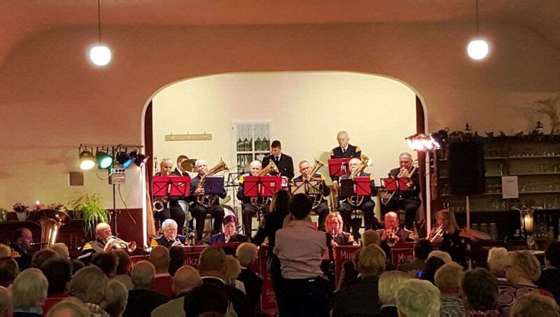 Ein Saal mit Publikum an Tischen. Im Hintergrund spielt der Musikzug unter einem Rundbogen.