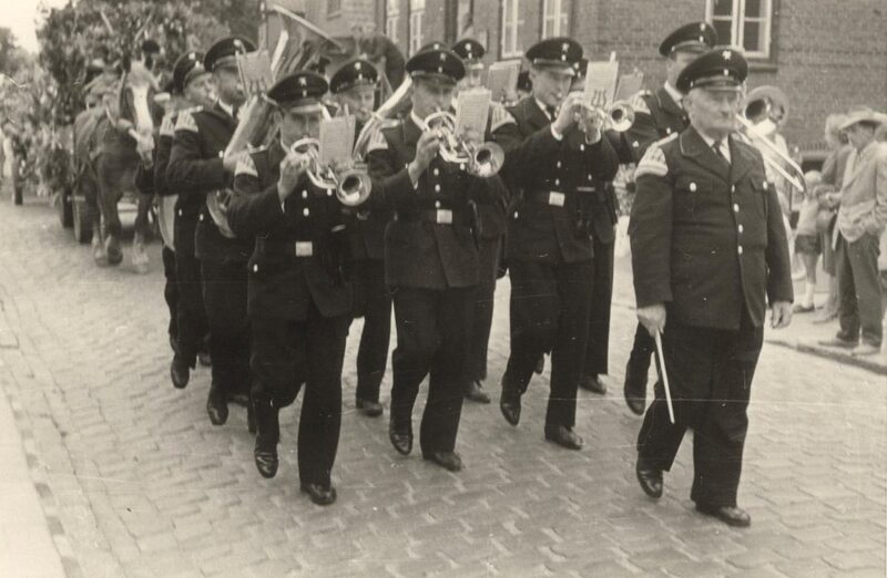 Schwarz-Weiß-Foto: Musiker in dunkler Uniform und Schirmmütze marschieren mit Trompeten und Hörnern über eine Dorfstraße.