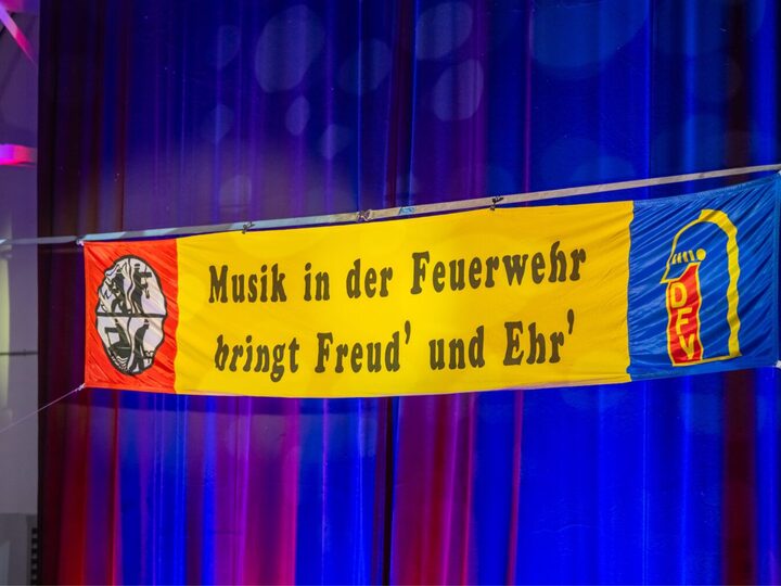 Ein gelbes Spruchband vor einem blau beleuchteten Bühnenvorhang: Musik in der Feuerwehr bringt Freud’ und Ehr’.