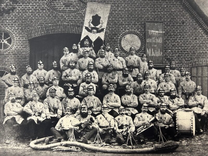 Altes Schwarz-Weiß-Foto: etwa fünfzig Feuerwehrmänner mit Helm und heller Uniformjacke in mehreren Reihen vor einem Backsteinhaus. Auf den Jacken stehen die Buchstaben K und F. In der Mitte eine Fahne, vorn ein Saugschlauch, rechts eine große Trommel.